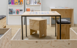 In der Leseecke sind alle Möbel aus hellem Holz. In der Mitte steht ein Tisch, auf dem ordentlich ein Memory ausgelegt ist, am Tisch stehen zwei Hocker. Im Hintergrund rechts steht ein weiterer Tisch, links lehnen zwei Bücherregale an der Wand, die bunten