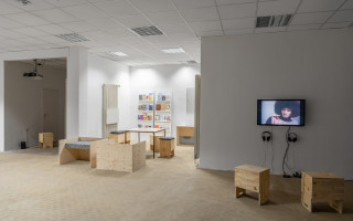 Im Ausschnitt aus dem Ausstellungsraum ist die Bücherecke zentral. Rechts hängt ein Monitor mit einem Videostill aus edna bonhommes Video „To Be Black, Free, and En(raged)“ (2020) an der Wand. Darunter hängen zwei Kopfhörer, davor stehen zwei Holzhocker. 